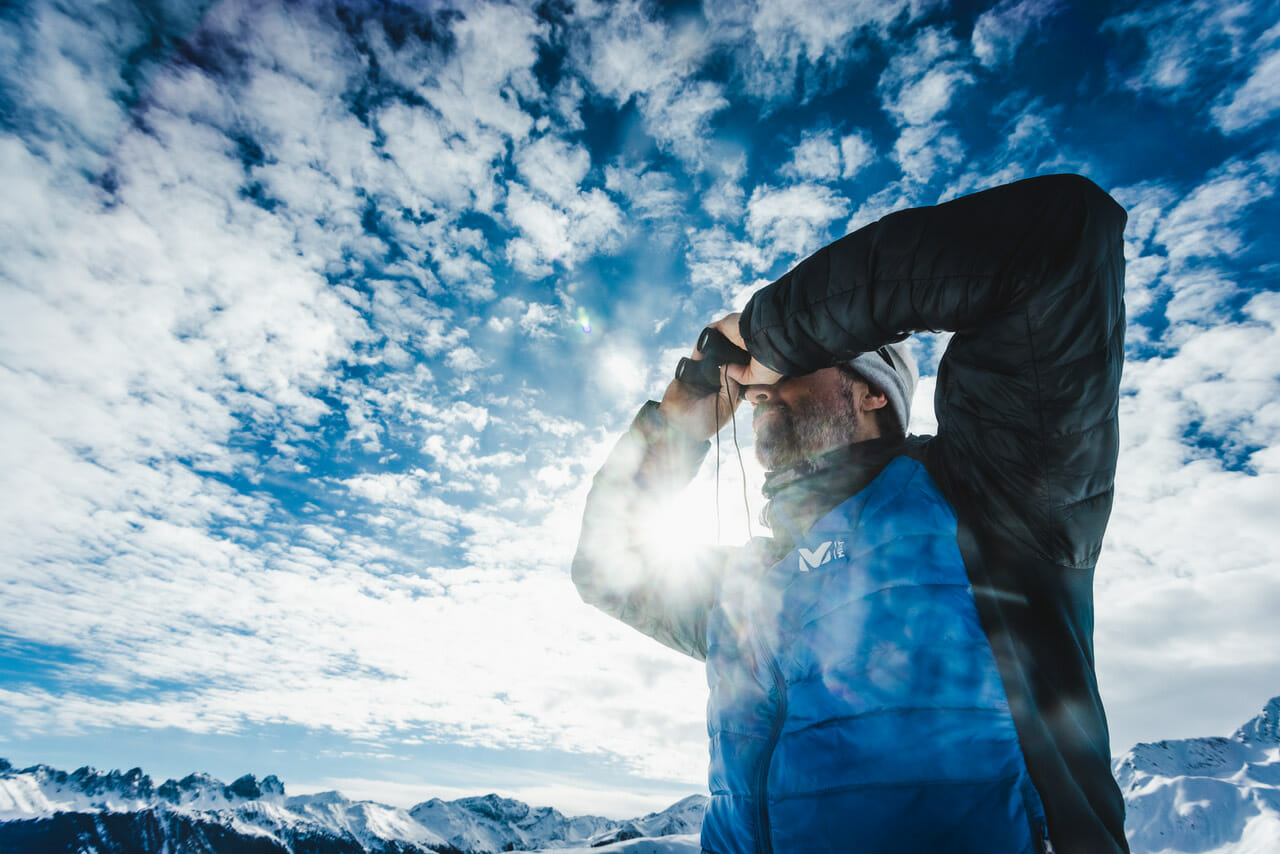 Stephan Görgl mit Feldstechern die Berge beobachtend, im Hintergrund Wolken,Berge, blauer Himmel