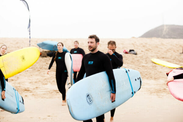 Gernot am Strand mit Surfboard unterm Arm, im Hintergrund andere Surfschüler