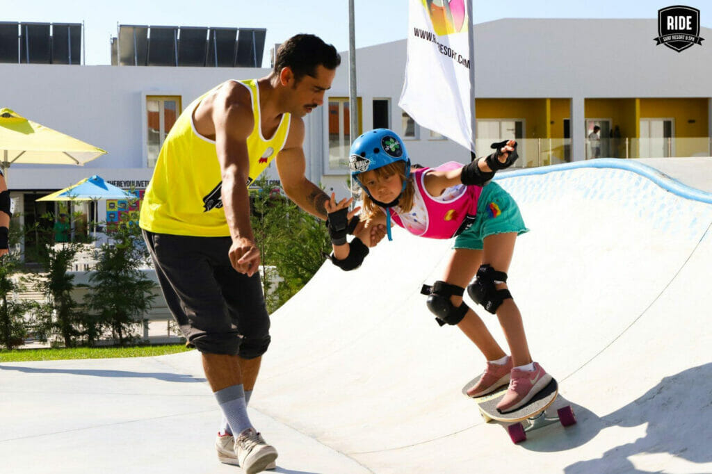 Mädchen auf Skatboard,Skatelehrer,Miniramp,Haus und bunter Schirm im Hintergrund